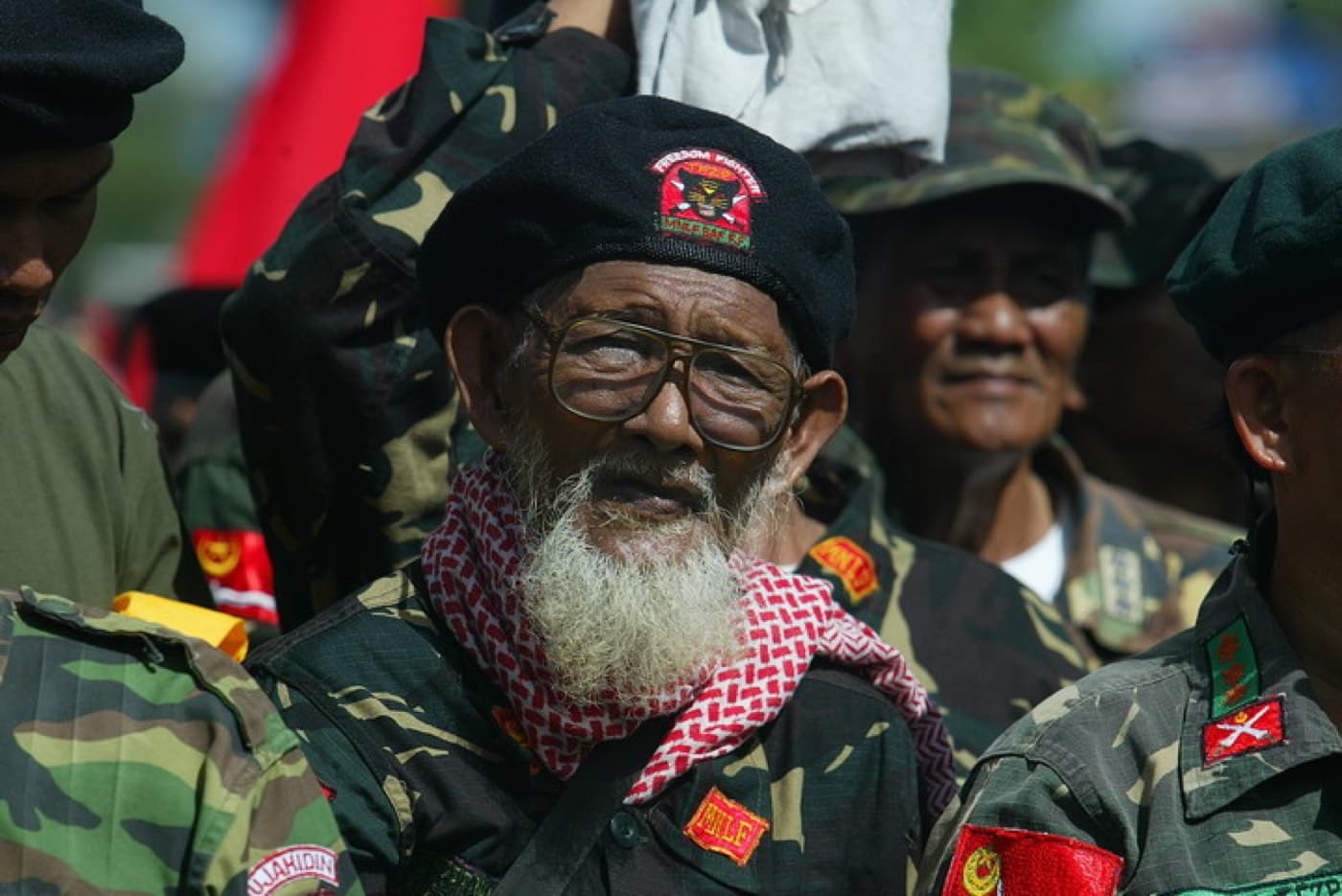 An elderly MNLF veteran in camouflage and a black beret bearing a Bangsa Moro badge, other fighters behind him.