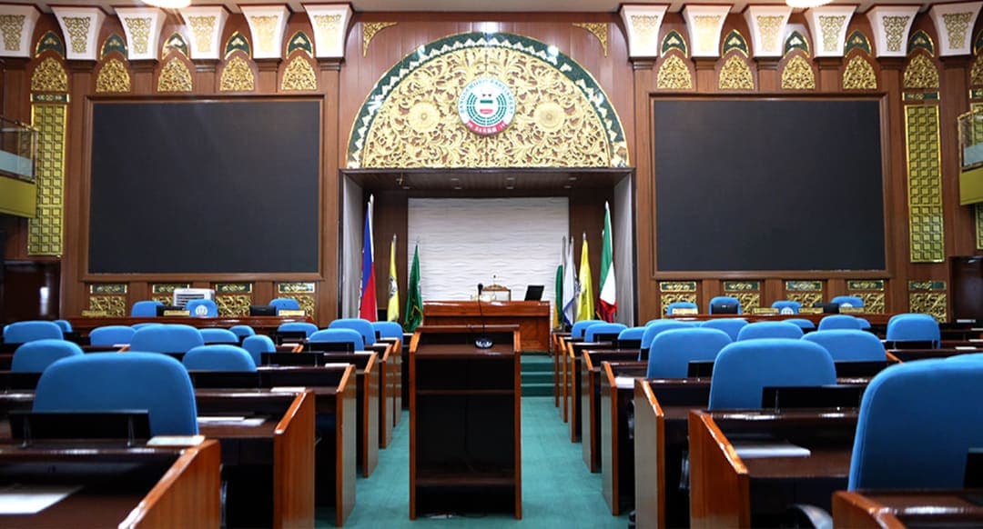 Rows of blue desks with monitors facing a green and gold rostrum in the Bangsamoro parliament session hall.