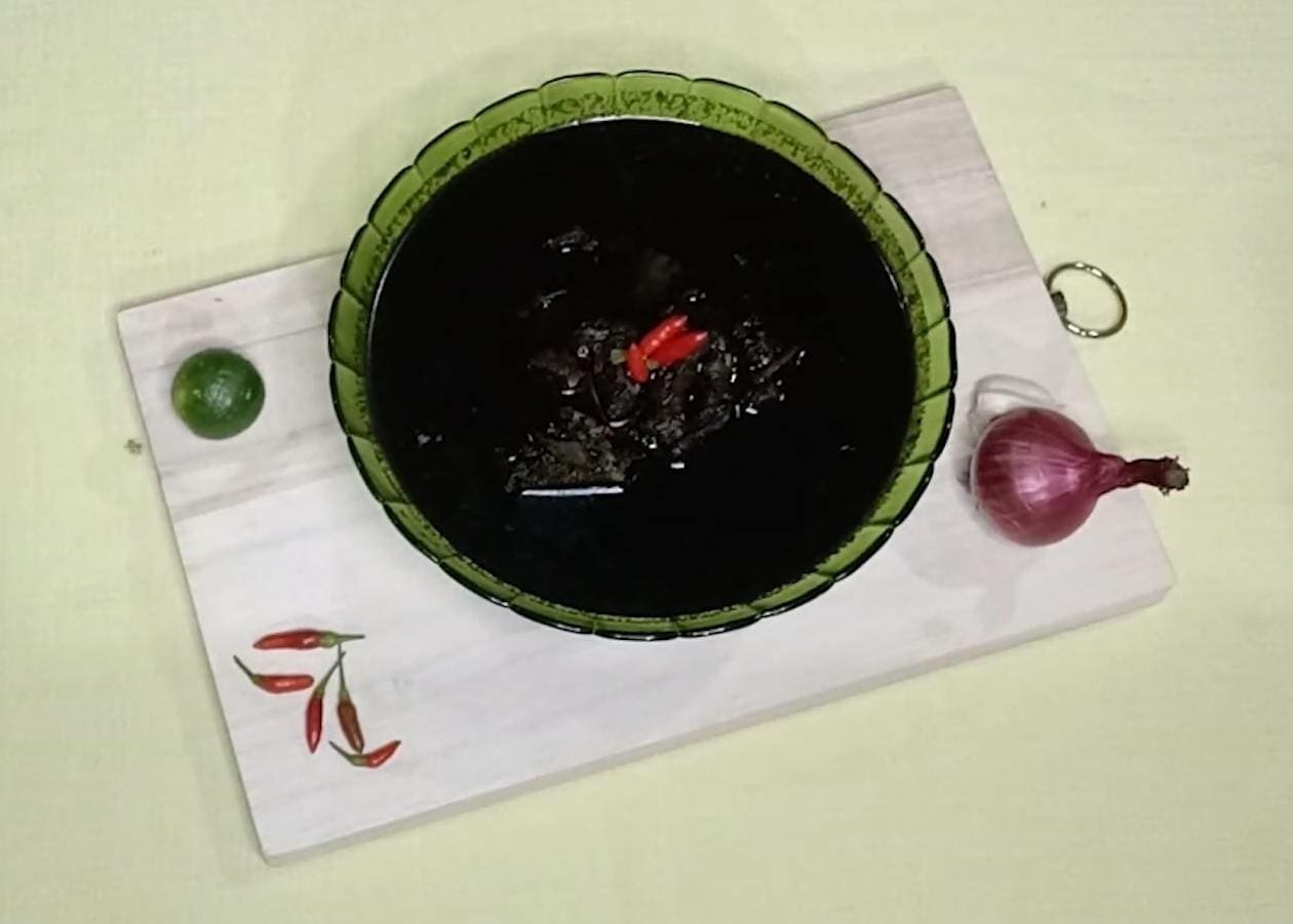 A bowl of near-black beef soup beside a red onion and dried chillies.